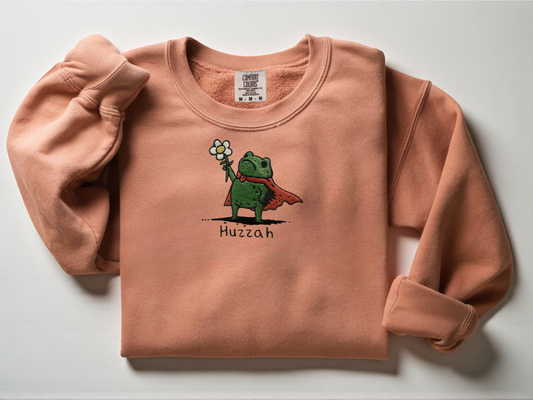 Close-up detail of embroidery stitching showing a cute frog knight holding a flower with the text Huzzah underneath on a Terracotta Comfort Color Crewneck.