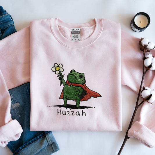 Pink sweatshirt with a green frog holding a flower and the word 'Huzzah' printed on it, placed on a white surface with denim jeans and cotton branches.