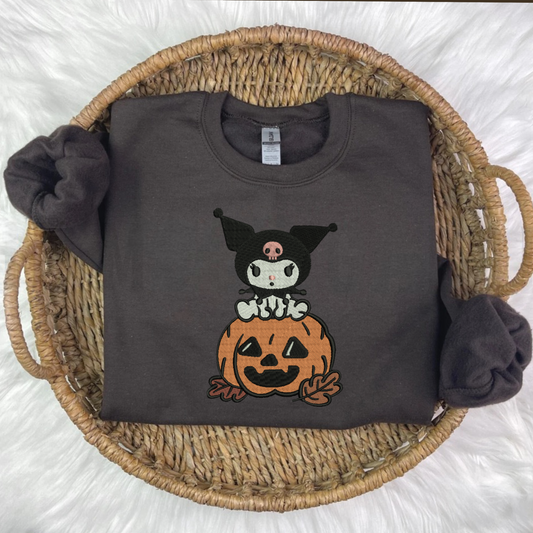 A black embroidered sweatshirt with a Kuromi and pumpkin embroidery, displayed on a surface with autumn decorations around it.