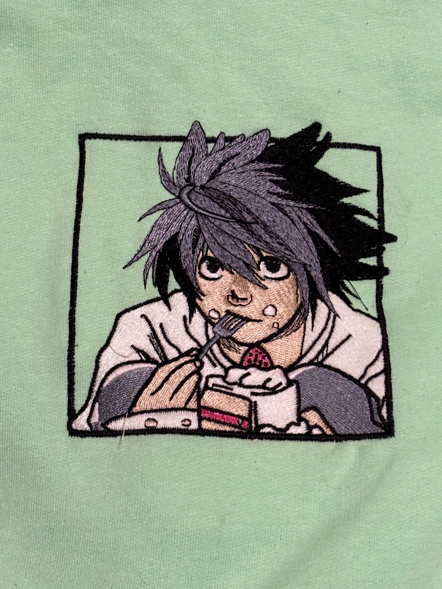 A character named L from the Anime Series, "Death Note' is on a green embroidered hoodie eating cake.