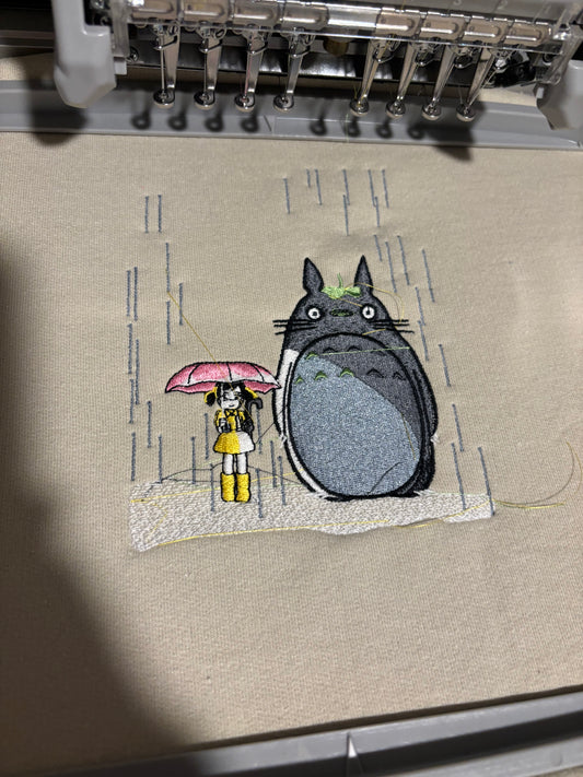 A beige embroidered sweatshirt with a scene from My Neighbor Totoro, held by a person with a pink umbrella in the background.