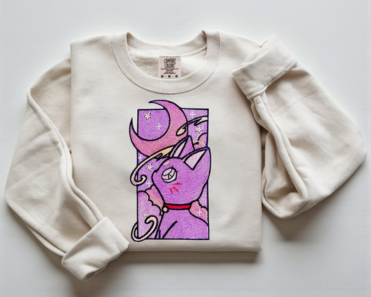 A White Comfort Colors Crewneck with an embroidered design of the Sailor Moon Cat with purple stitching.
