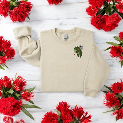 Embroidered crewneck sweatshirt with a graphic of a green frog holding a bow and arrows, and a heart-shaped balloon, against a floral background.