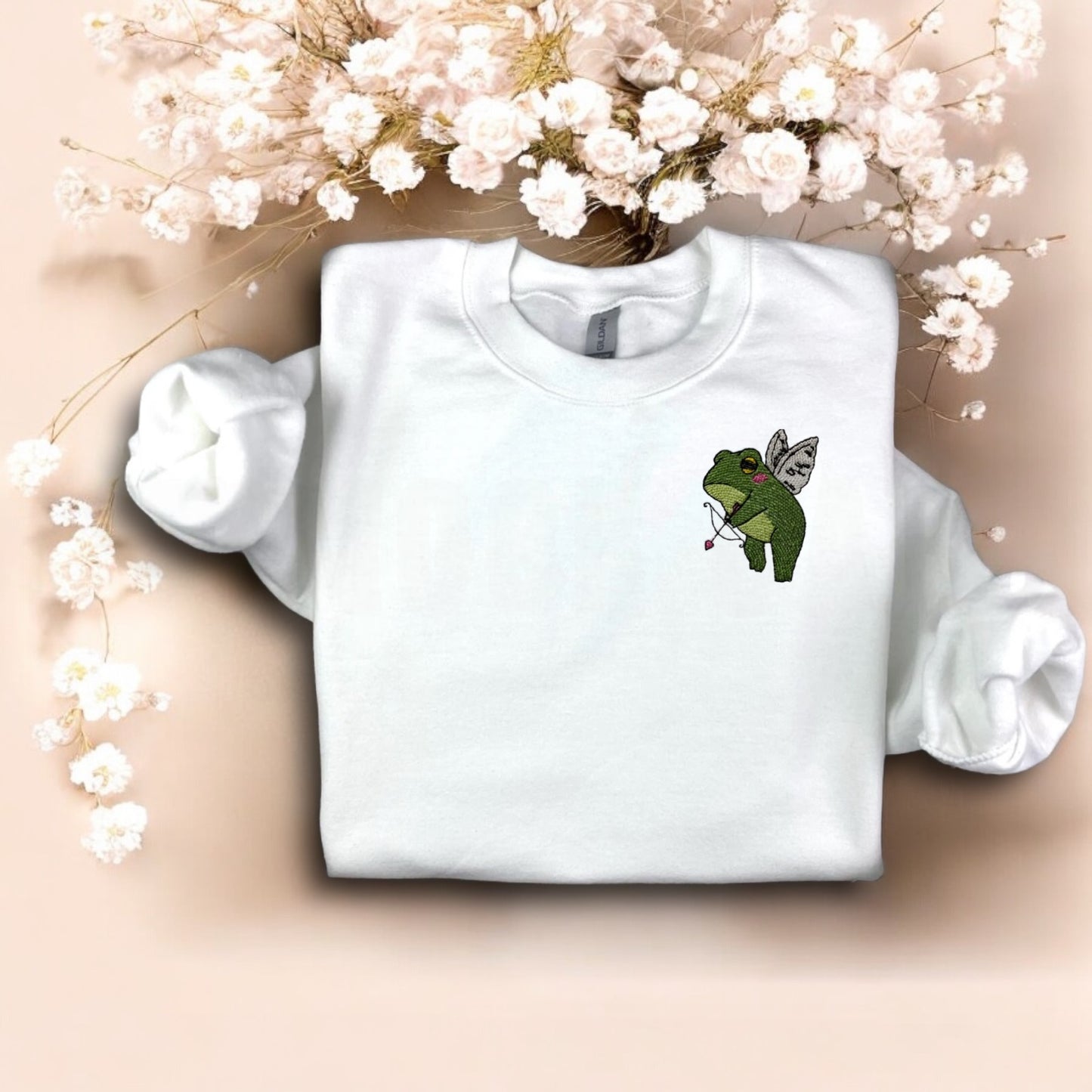 A white crewneck sweatshirt with a graphic of a green frog holding a bow and arrows, and a heart-shaped balloon, against a floral background.