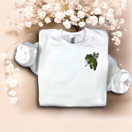 A white crewneck sweatshirt with a graphic of a green frog holding a bow and arrows, and a heart-shaped balloon, against a floral background.
