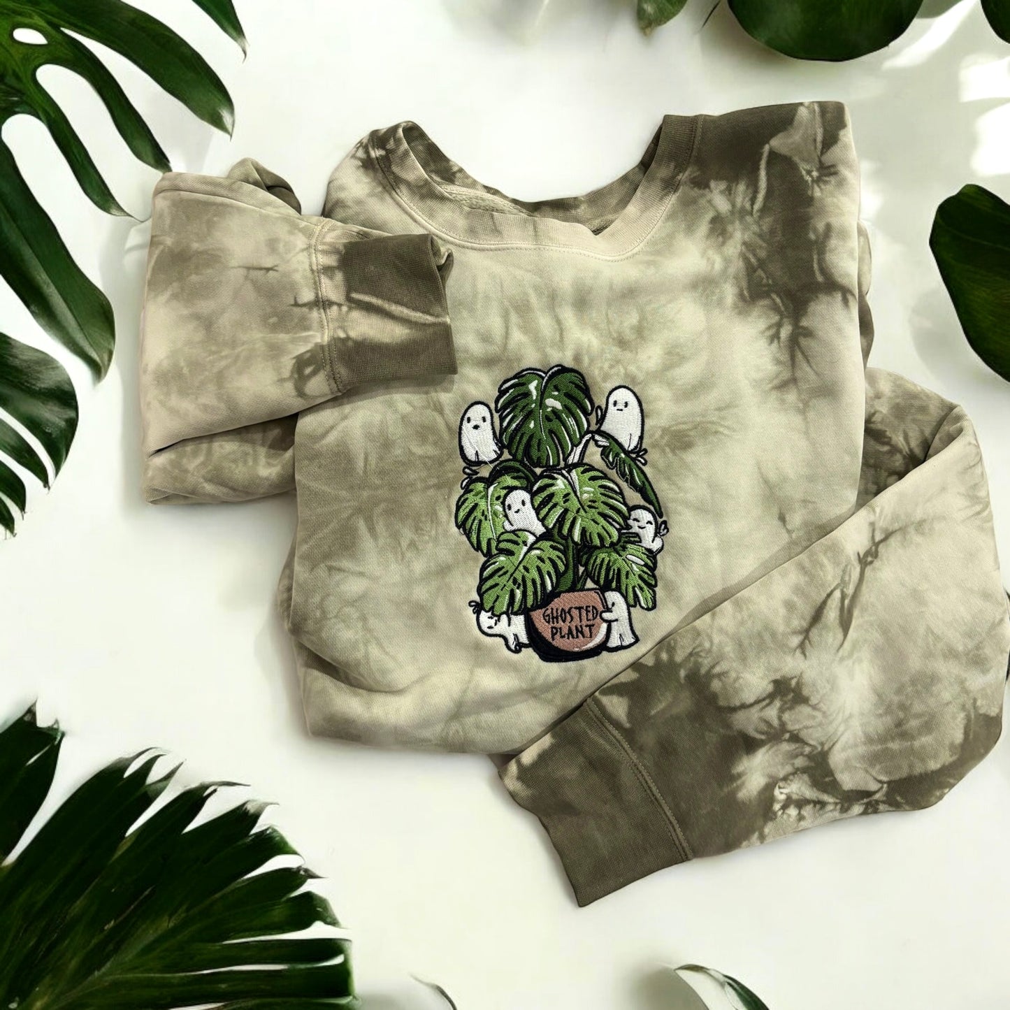 An Olive tie-dye sweatshirt with a front graphic of ghosts and a plant, displayed on a flat surface with a green plant in the background.