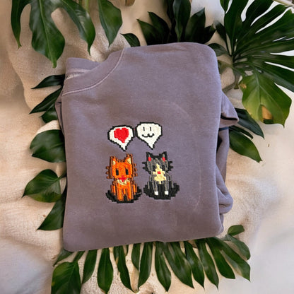 A purple embroidered sweatshirt with a graphic of two cartoon cats and the text 'Stardew Kitty Friends' on it, placed on a flat surface with a green plant in the background.