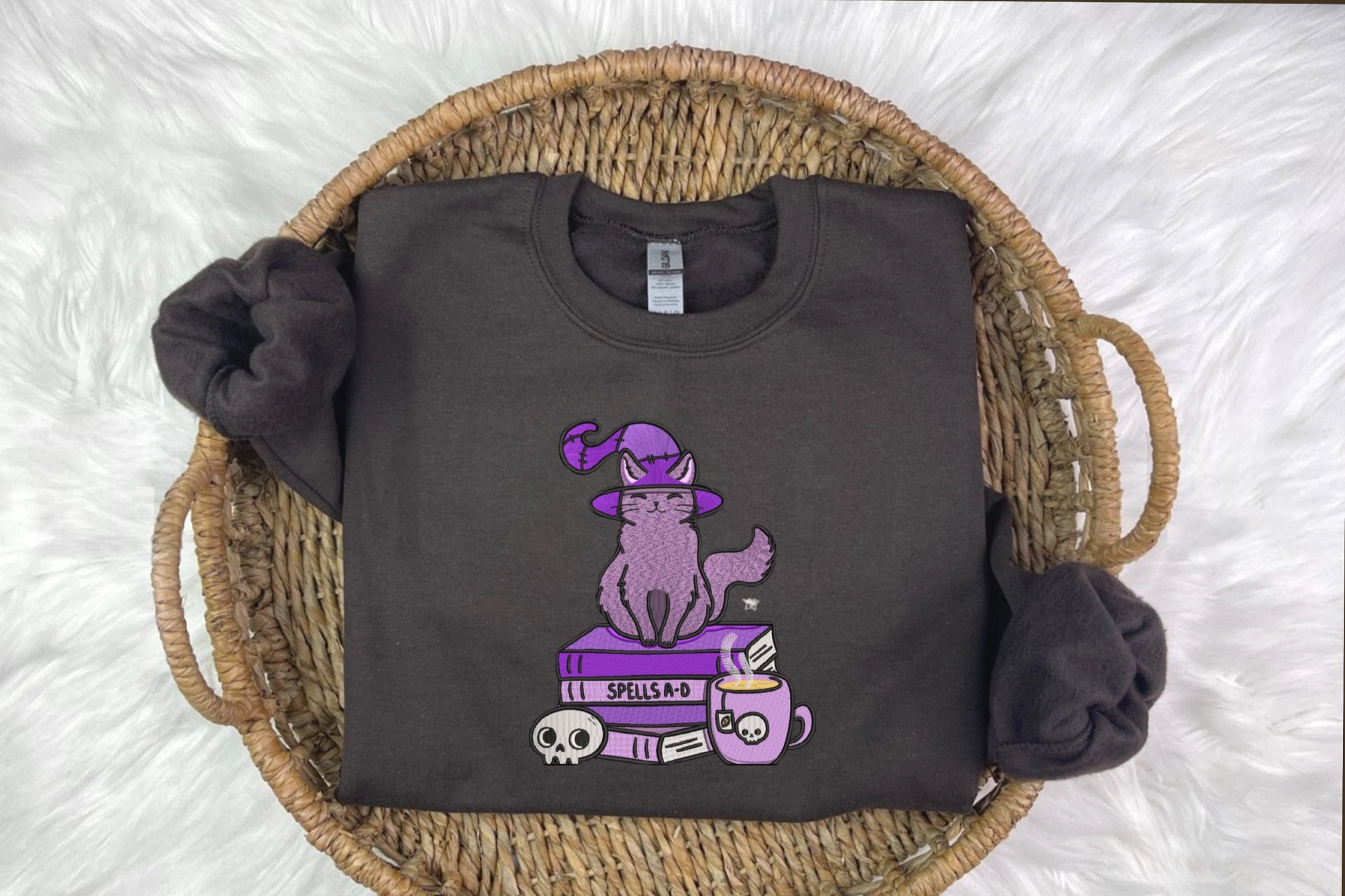 A black embroidered sweatshirt with a design of a cat wearing a witch's hat, perched on a book, presented on a woven basket background.