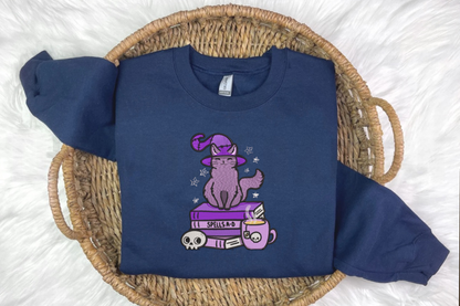 A blue embroidered sweatshirt with a design of a cat wearing a witch's hat, perched on a book, presented on a woven basket background 