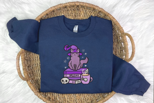 A blue embroidered sweatshirt with a design of a cat wearing a witch's hat, perched on a book, presented on a woven basket background 