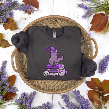 A black embroidered sweatshirt with a design of a cat wearing a witch's hat, perched on a book, presented on a woven basket background with purple flowers scattered around.