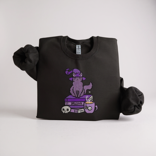 A black embroidered sweatshirt with a design of a cat wearing a witch's hat, perched on a book, presented on a woven basket background.