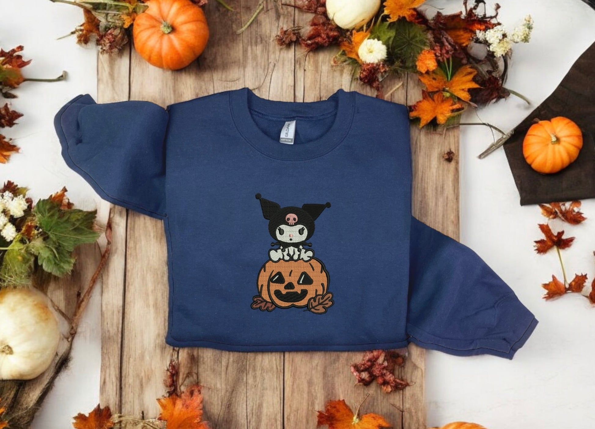 A navy blue sweatshirt with a Kuromi and pumpkin embroidery, displayed on a surface with autumn decorations around it.