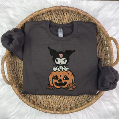 A black embroidered sweatshirt with a Kuromi and pumpkin embroidery, displayed on a surface with autumn decorations around it.