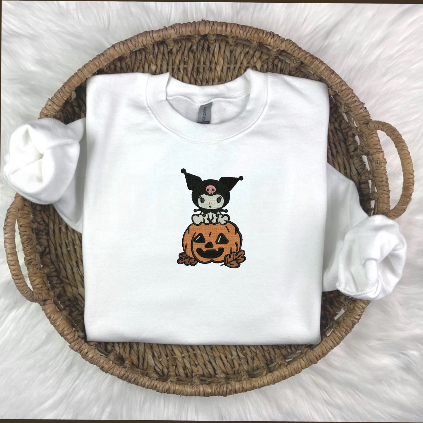 A white embroidered sweatshirt with a Kuromi and pumpkin embroidery, displayed on a surface with autumn decorations around it.