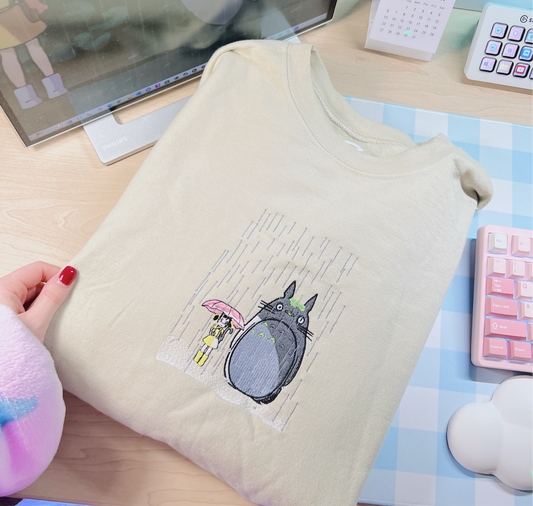 A beige embroidered sweatshirt with a scene from My Neighbor Totoro, held by a person with a pink umbrella in the background. There is a computer monitor, keyboard, and other anime-themed accessories in the background.