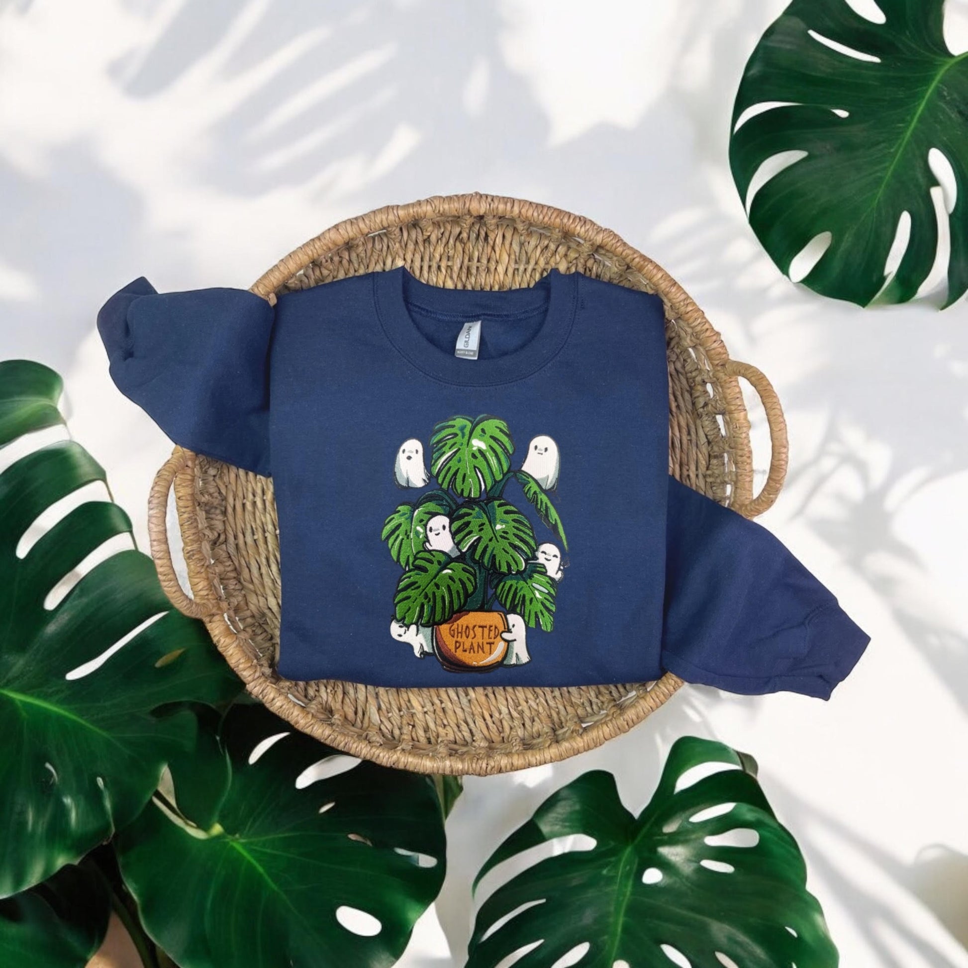 A navy embroidered sweatshirt with a front graphic of ghosts and a plant, displayed on a flat surface with a green plant in the background.