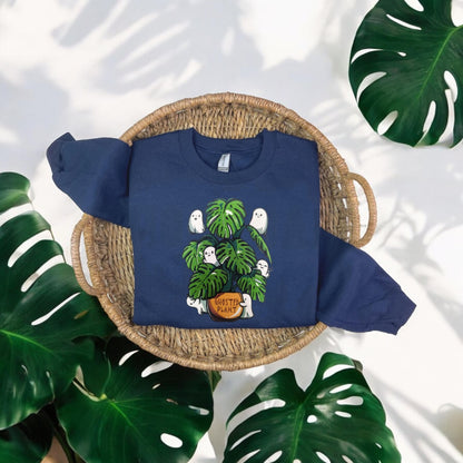 A navy embroidered sweatshirt with a front graphic of ghosts and a plant, displayed on a flat surface with a green plant in the background.