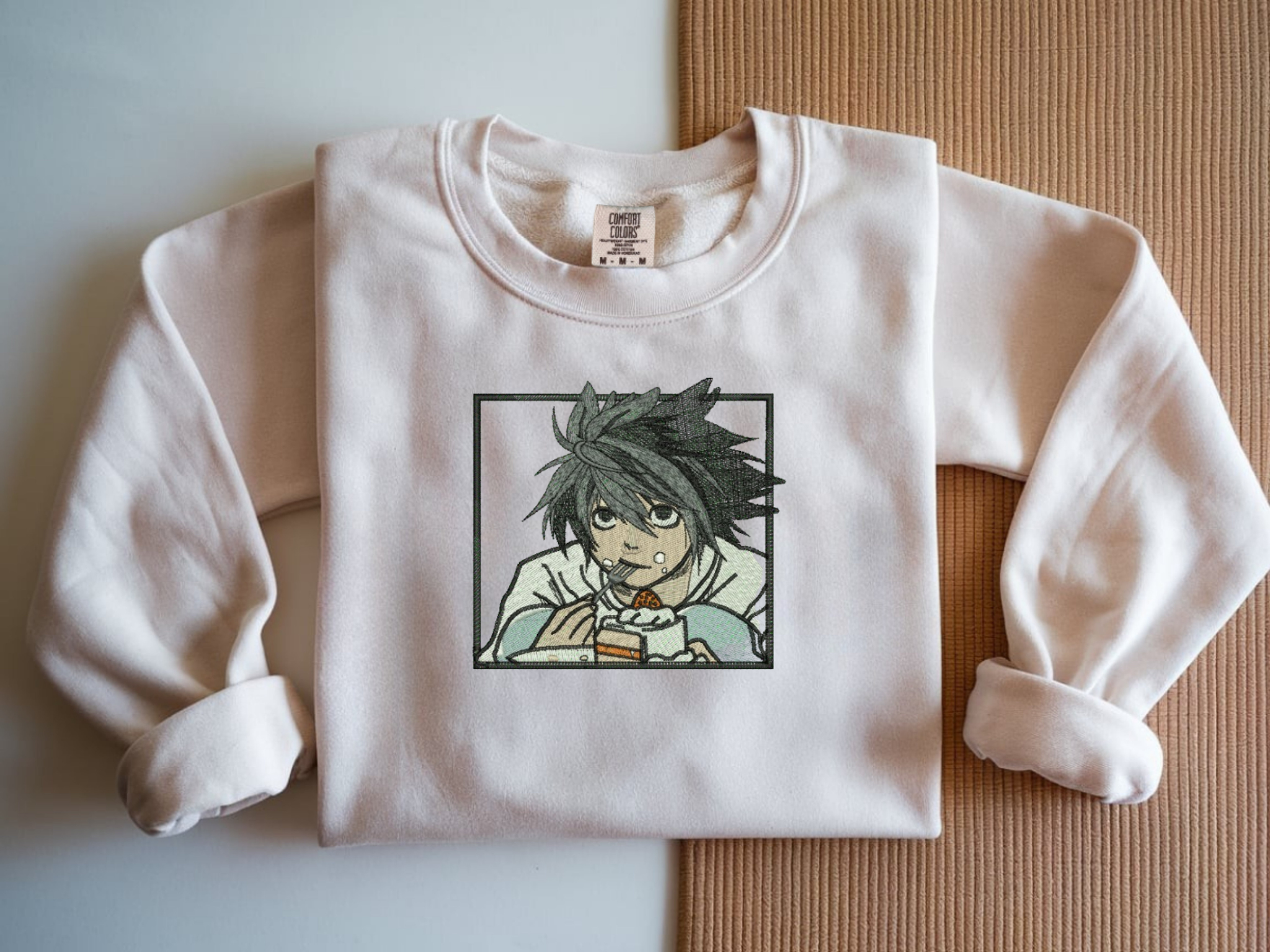 White Comfort Colors crewneck sweatshirt with minimalist black line embroidery of L from Death Note crouching eating cake.