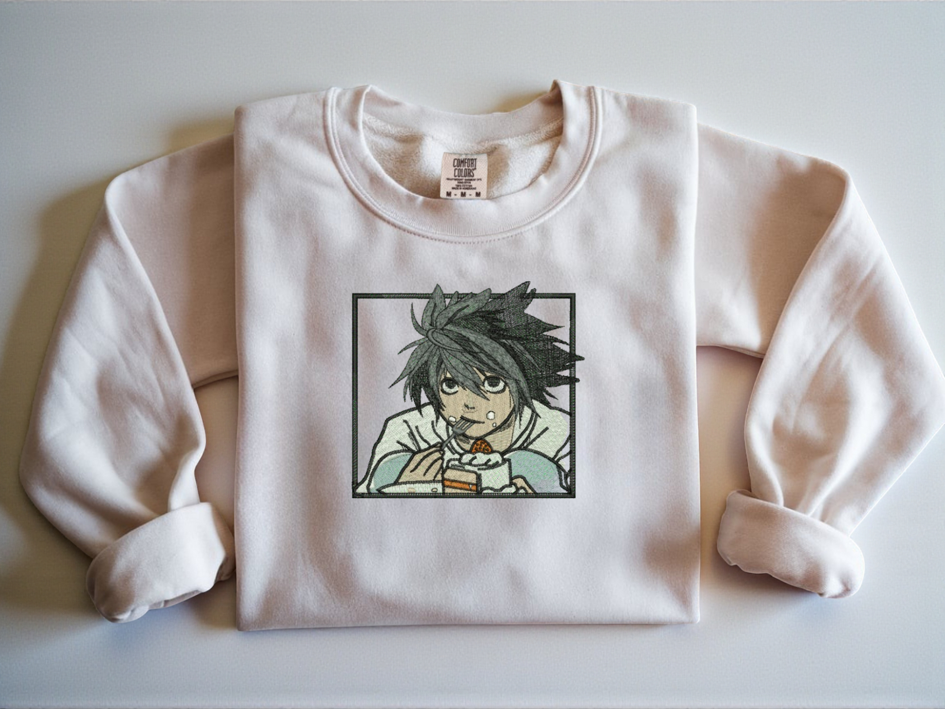 White Comfort Colors crewneck sweatshirt with minimalist black line embroidery of L from Death Note crouching eating cake.